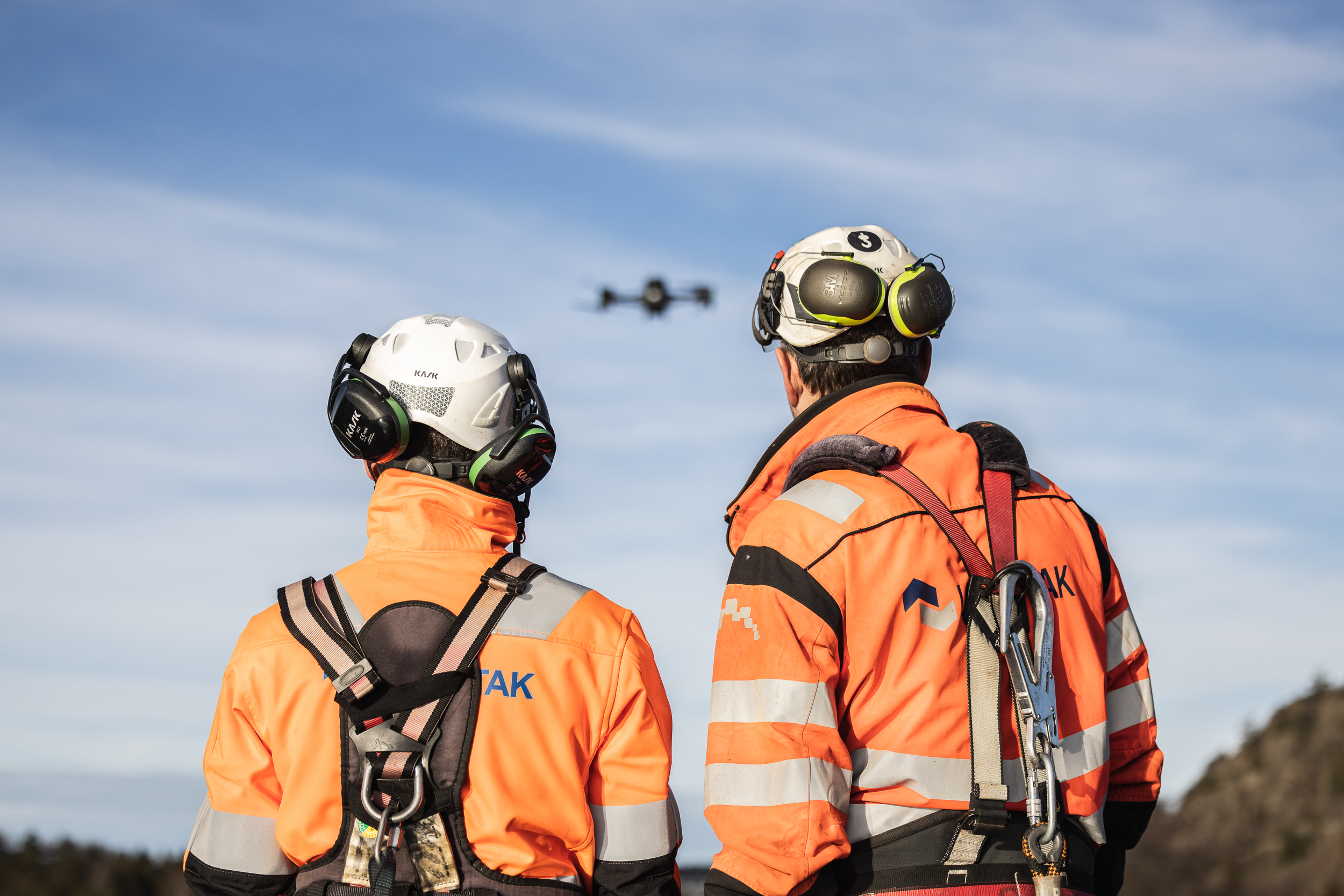 To takarbeidere står med ryggen til og ser på en drone i luften